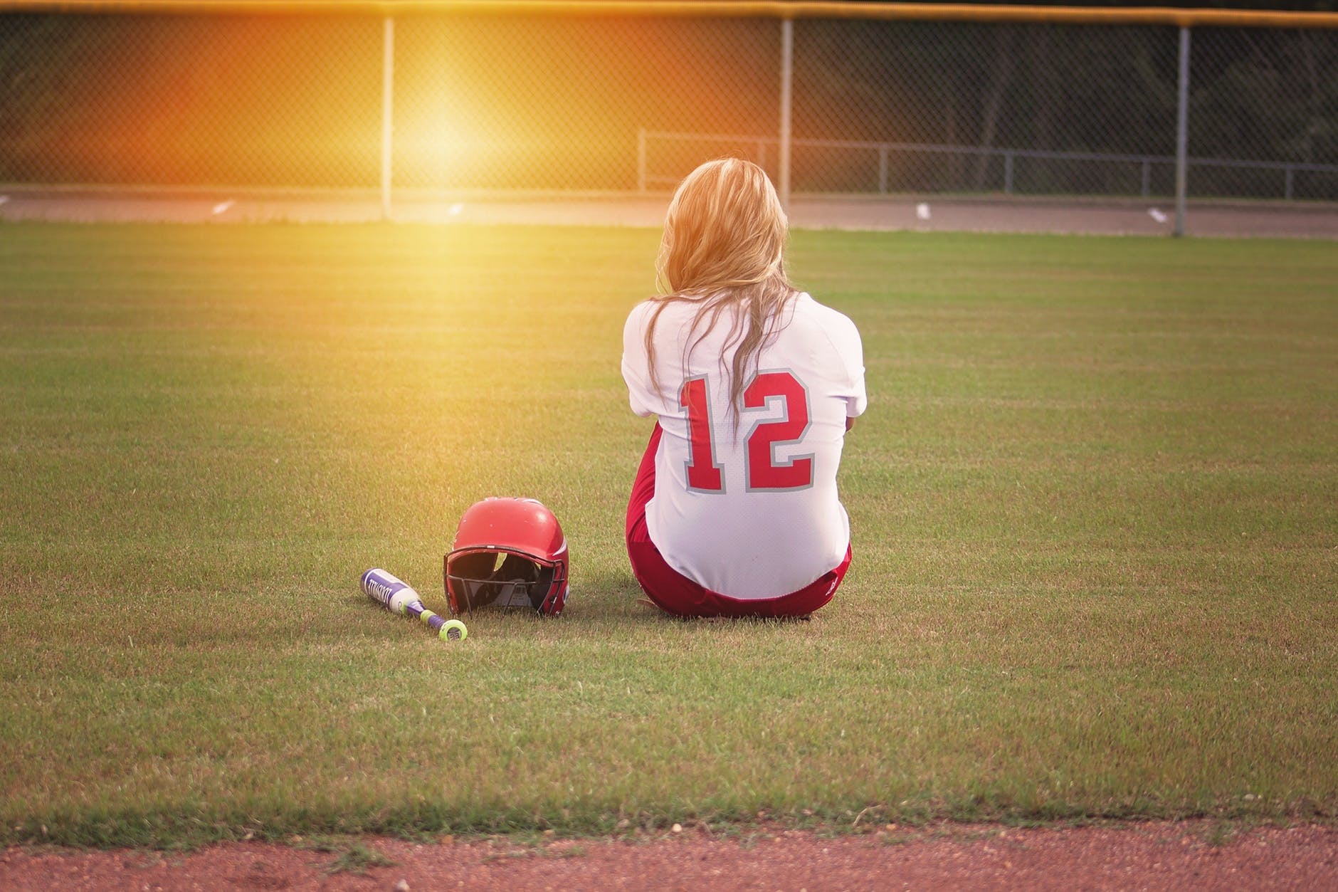female baseball player sitting on grass field beside helmet and baseball bat